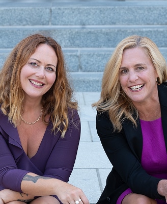 Katie and Jennifer smiling while seated on stone steps, dressed in coordinating purple tones, conveying connection, confidence, and partnership