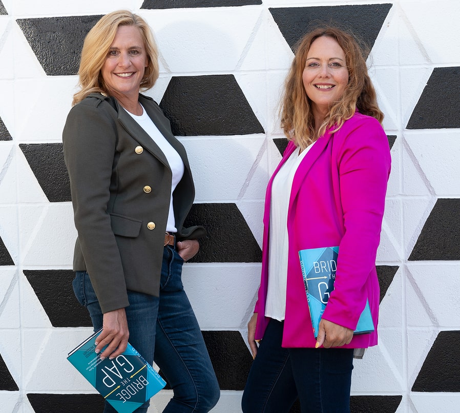 Jennifer and Katie standing against a geometric black-and-white wall, smiling confidently while holding their book Bridge the Gap and wearing blazers with jeans